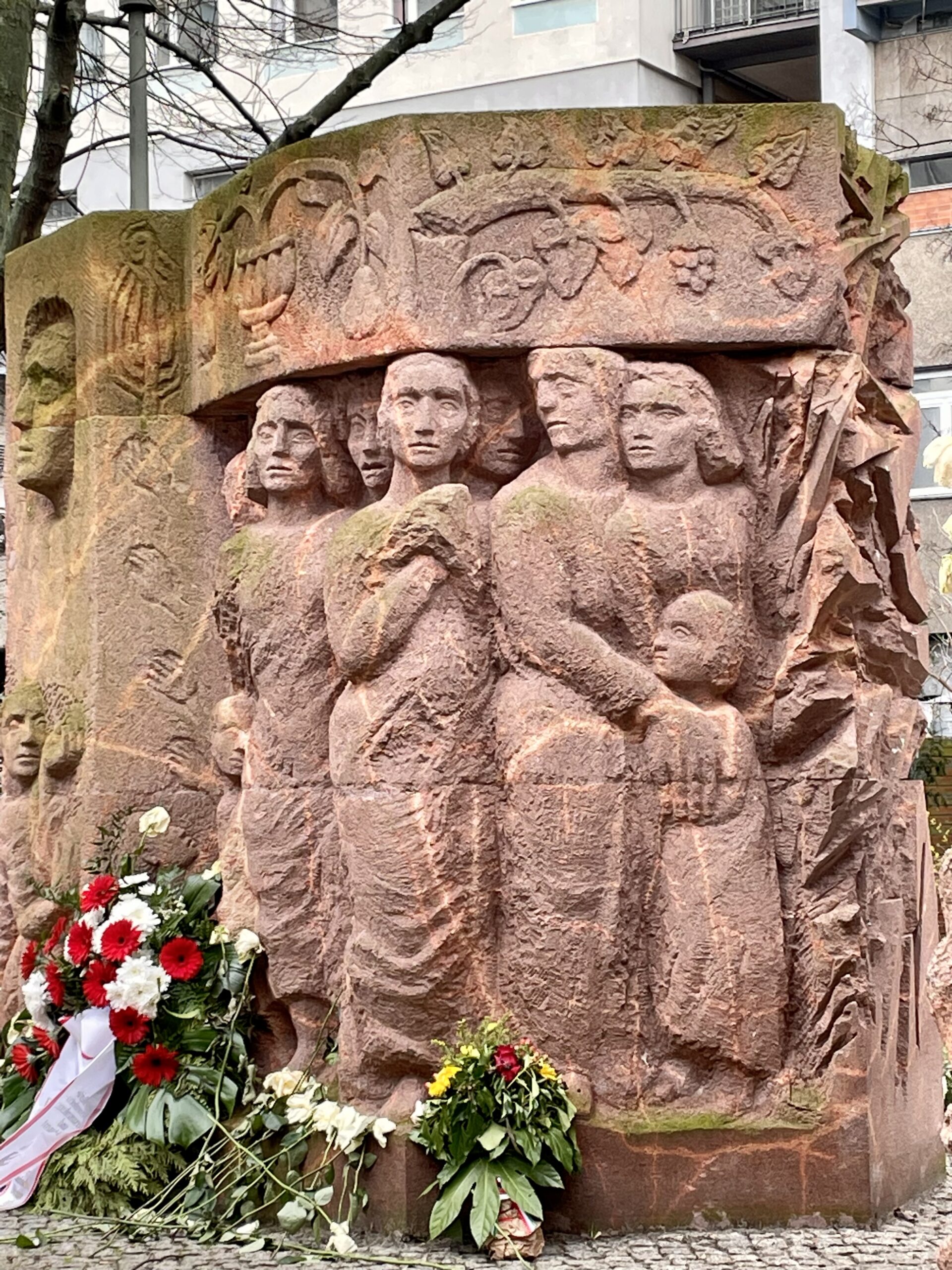 Eine rotfarbene Steinskulptur mit Gesichtern und Menschen drauf. Davor liegen rote, weiße und gelbe Blumen und ein Blumenkranz.