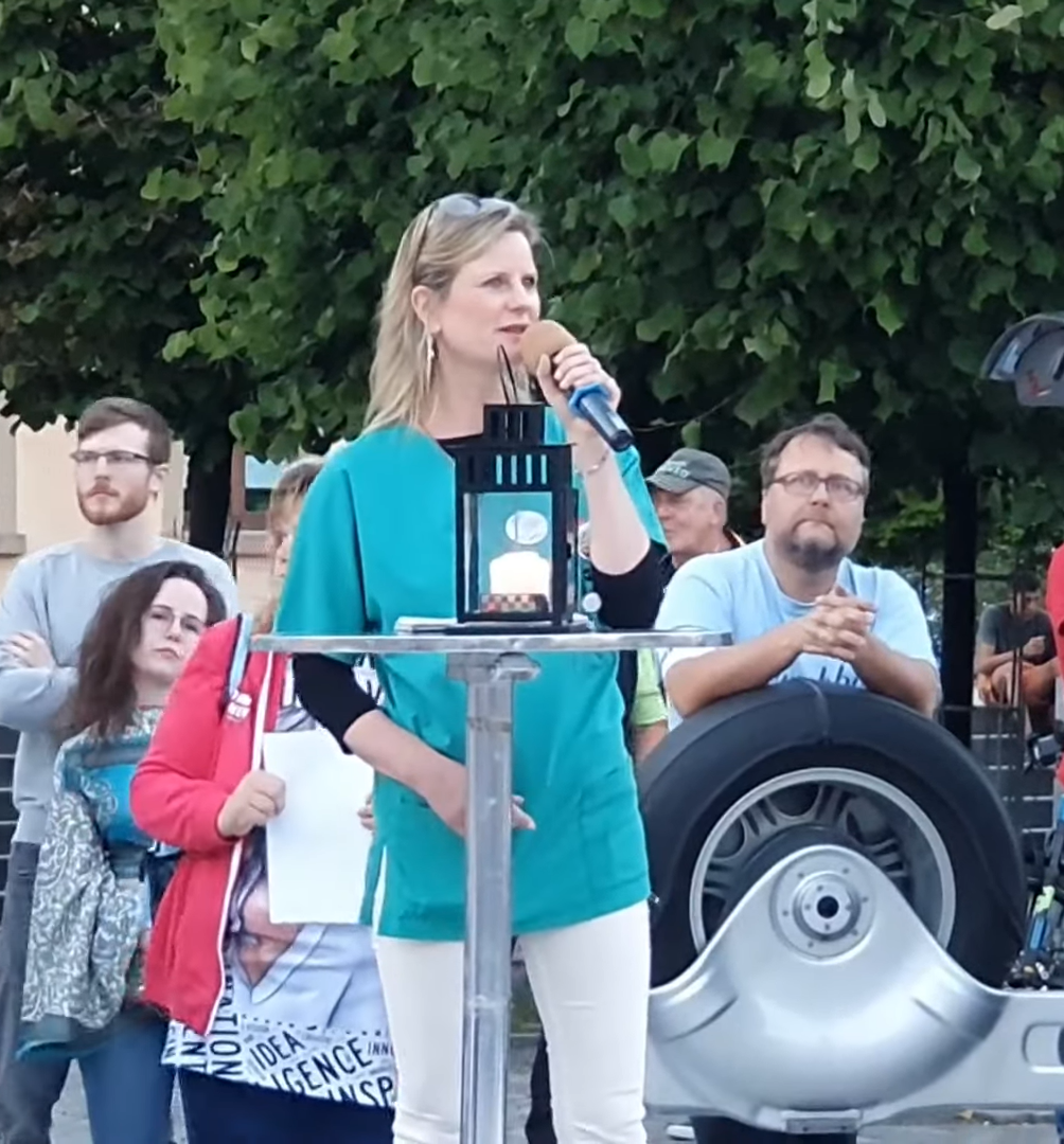 Eine Frau mit blauem Oberteil und weißer Hose steht vor einer Gruppe von Menschen mit einem Mikfrofon. Vor ihr ist ein silbener Stehtisch mit einer schwarzen Sockelleuchte.