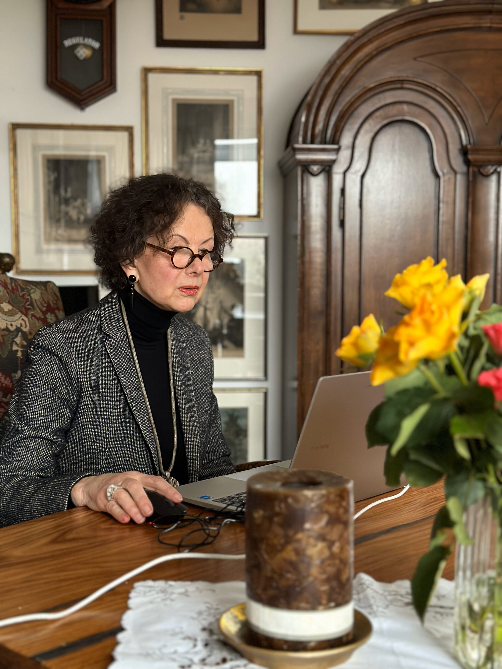 Eine Frau mit braunen, lockigen Haaren und schwarzer Brille sitzt vor einem Laptop. Der Tisch ist mit einer Kerze und Blumen geschmückt. Hinter ihr antike Möbel.