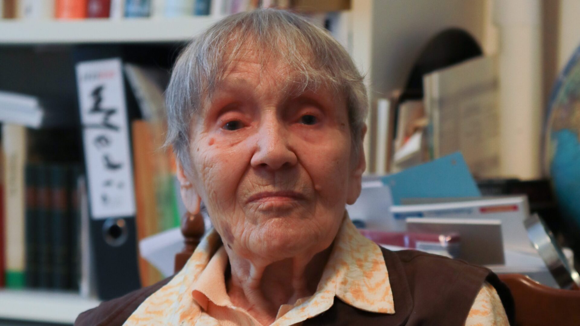 Eine ältere Dame mit kurzen, grauen Haaren und einer orangenen Bluse sitzt vor einem vollen Bücherregal. Sie blickt in die Kamera.