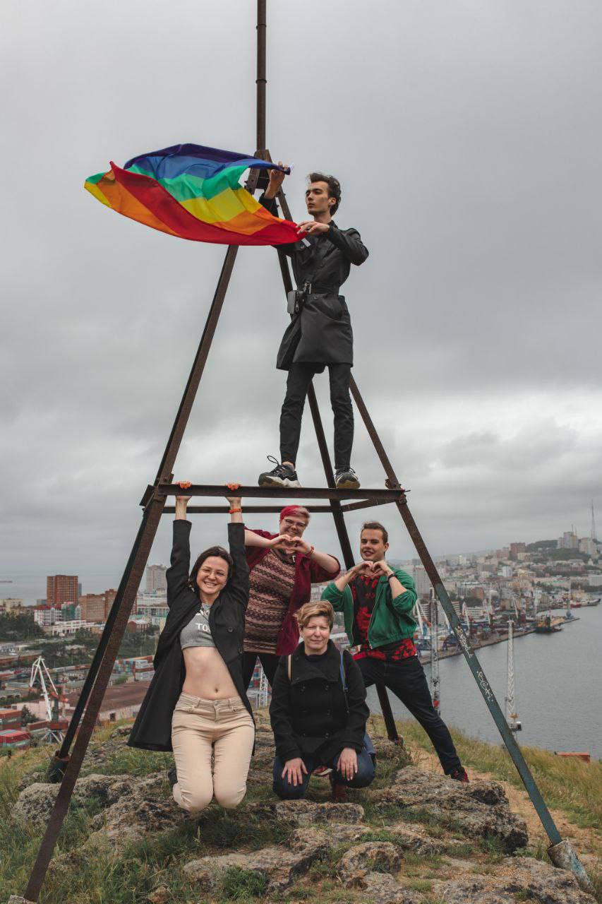 Fünf junge Menschen posieren an einem pyramidenförmigen Drahtgestell. Es liegt auf einem Hügel vor einem Hafen. Einer der Jugendlichen in schwarzer Lederjacke steht auf dem Gestell und hält eine Regenbogenfahne in den Händen.