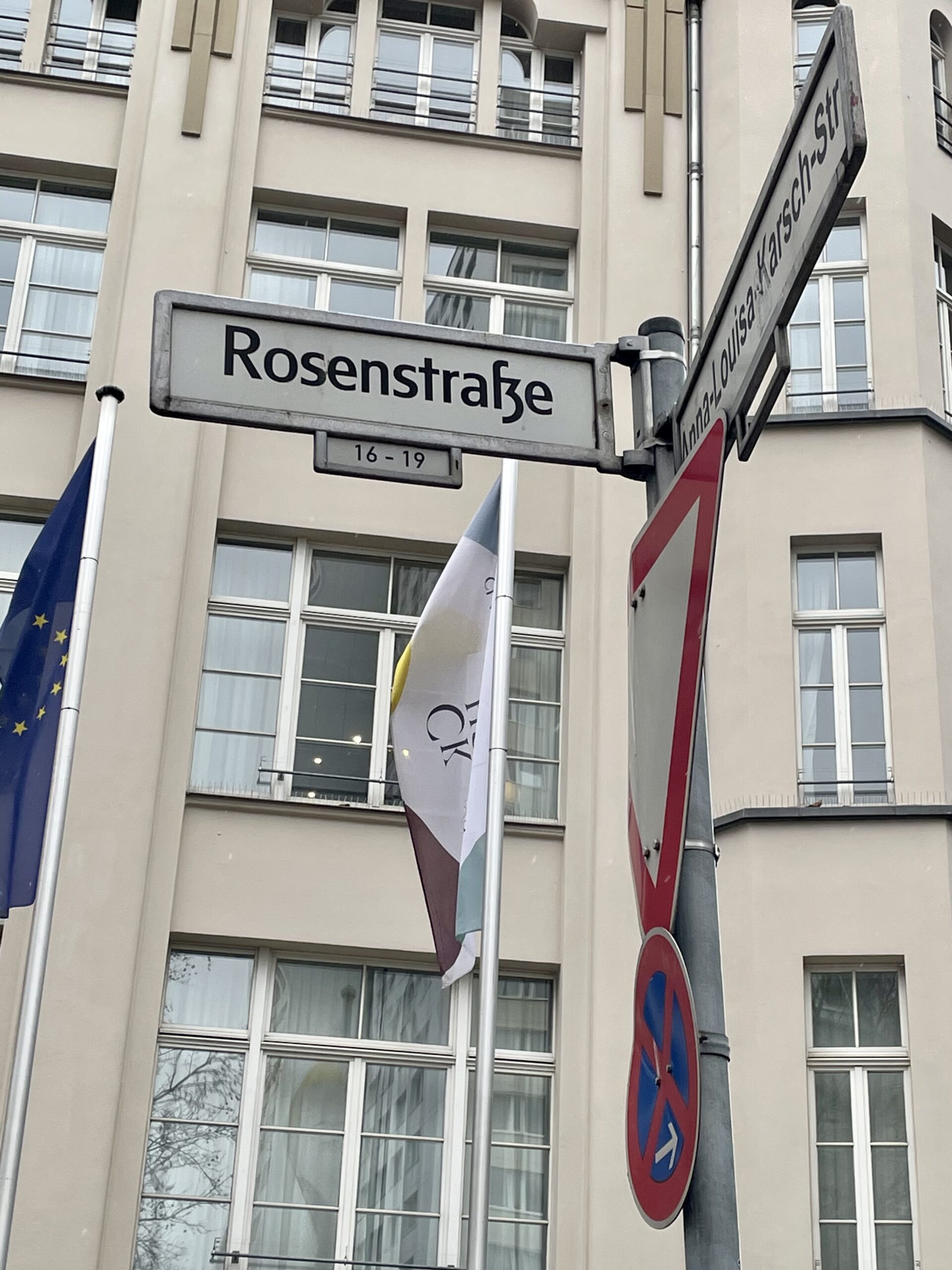 Vor einem beigen Gebäude mit großen Fenstern steht ein Mast. Daran sind Verkehrs- und Straßenschilder. Auf dem Straßenschild steht: Rosenstraße 16-19.